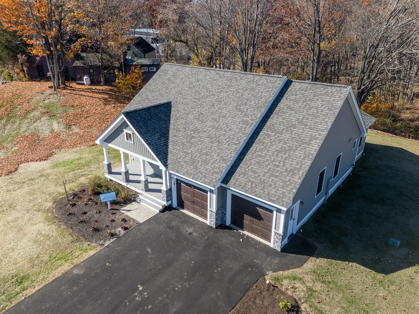 Aerial real-estate photograph of a contemporary New England home with white trim, gray shingle roof, and a double garage on a wooded lot — captured for Britten Aerial Services